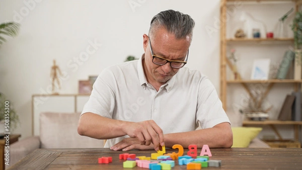 Obraz Korean male pensioner with Alzheimer’s disease trying to improve memory by doing rehab exercise for brain at home. He murmurs to himself while looking and counting the letter blocks
