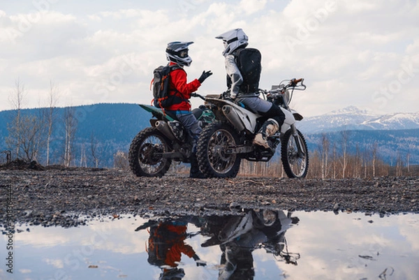 Fototapeta Females wearing helmets and motorcycle gear sitting on motocross motorcycles discuss the journey. Reflection in puddles, snowy peaks on horizon. Offroad travel