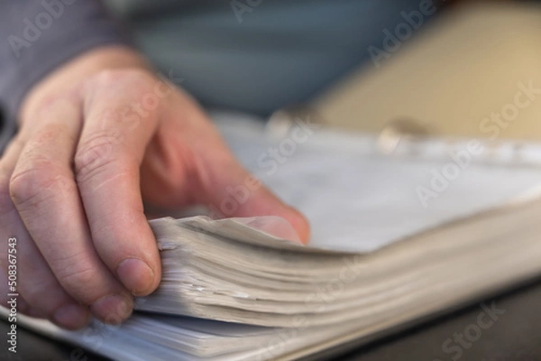 Obraz A close-up of a man's hand flipping through documents in a folder. An adult male is holding a report in an open gray folder with a binder. Selective Focus.