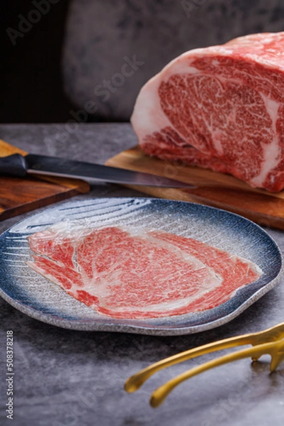 Fototapeta Premium Rare Slices sirloin Wagyu A5 beef with high-marbling texture pick up by bbq tongs with hand from stone plate. Served for Yakiniku, Sukiyaki and Shabu.