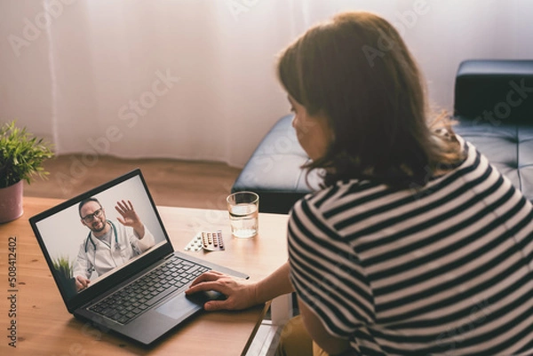 Obraz Woman sitting on a sofa and talking with a doctor online using laptop. Telemedicine concept.