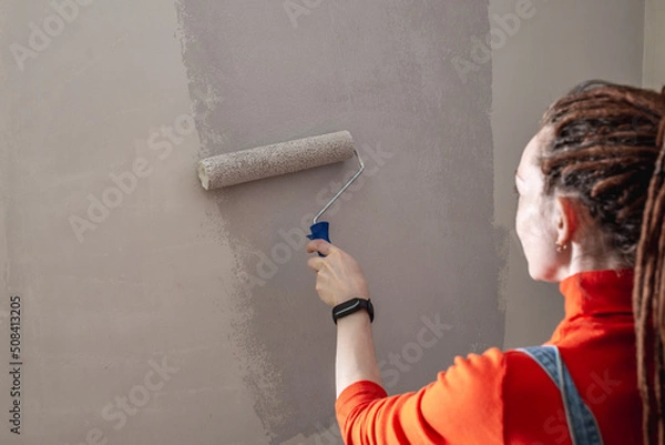 Obraz Woman with dreadlocks, in an orange turtleneck and denim jumpsuit is painting the wall with a roller in gray color. Concept of repair, renovation of the new apartment