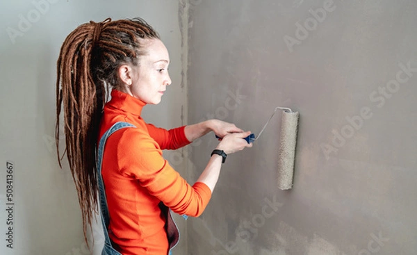 Obraz Woman with dreadlocks, in an orange turtleneck and denim jumpsuit is painting the wall with a roller in gray color. Concept of repair, renovation of the new apartment