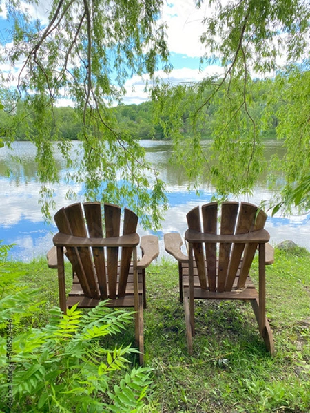 Fototapeta Chaises Adirondack en bois dans un jardin sur le bord de l'eau. Paysage d'été au travers les feuilles vertes retombantes d'un arbre. Chaise de patio sous un Saule pleureur