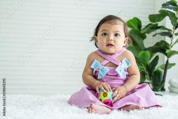 Fototapeta Adorable mixed race baby girl in pink clothes sitting on bed at home, looking away, playing toy color ball. Kid at home and childhood concept