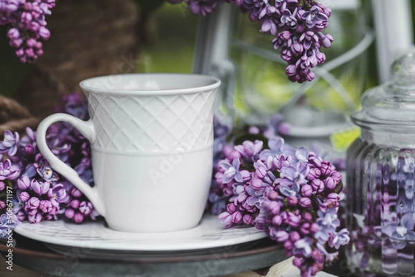 Obraz A beautiful postcard. A white coffee cup with a saucer, candles and a vase with a bouquet of purple lilac. The color 2022 is very peri. Beautiful still life. Spring time. The concept of "Good morning"