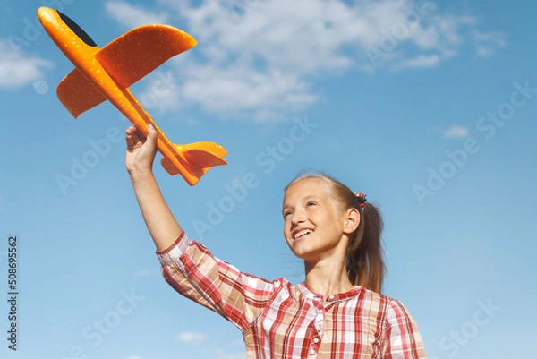 Obraz A girl with a toy airplane in her hand against a blue sky background. Happy childhood. Summer vacation. Journeys.