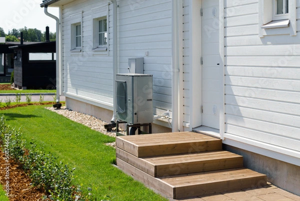 Fototapeta Air source heat pump installed on the exterior of a modern wooden house. Air source heat pump reduces heating costs and your home's carbon footprint.