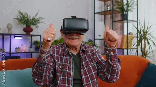 Fototapeta Senior elderly man using headset helmet app to play simulation game, simulation glasses gadget technology. Watching virtual reality 3D 360 video. Mature grandfather in VR goggles on couch at home
