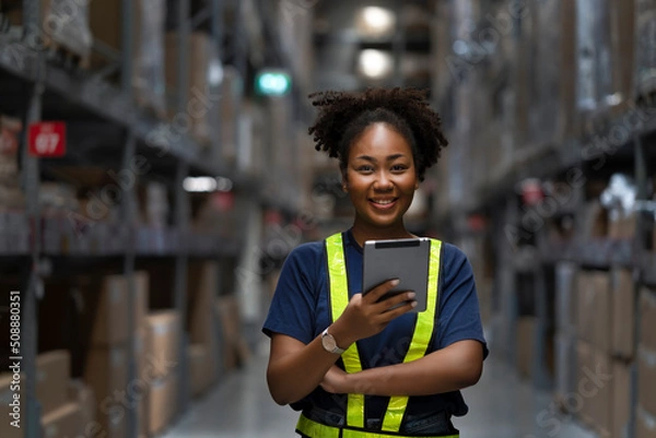 Fototapeta Young smart African American woman working in warehouse using the tablet for stock checking and inspection the boxes, logistic warehouse to deliver the shipment, happiness worker with smiling face