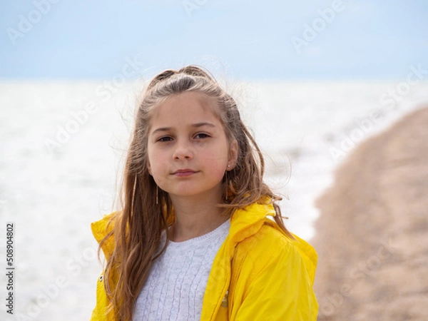Obraz A pretty girl with long hair stands against the background of the water surface.  Open portrait of a smiling blonde teenage girl in a yellow windbreaker on the seashore