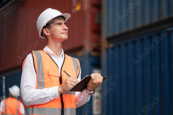 Obraz Dock manager or engineer worker in safety vest standing in shipping container yard holding tablet with smile. Import and export product. Manufacturing transportation and global business concept.