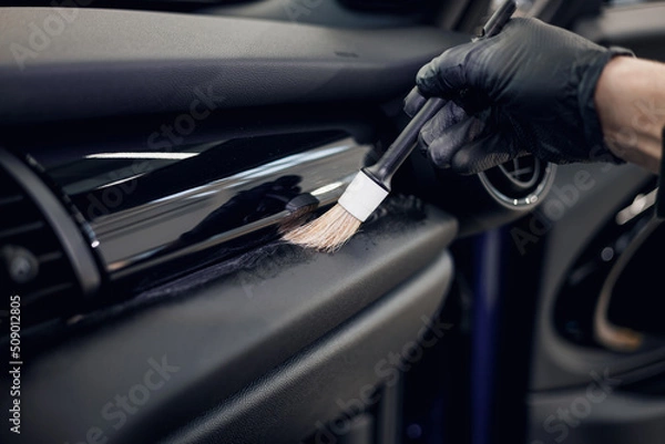 Obraz Cleaning the car interior with a brush. Auto detailing worker cleaning car interior, car detailing concept. Selective focus. Dust removal from the dashboard of the car with a brush