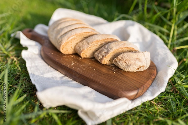 Fototapeta Composition of homemade white flour bread on wood board with white cloth napkin on the green grass . High quality photo