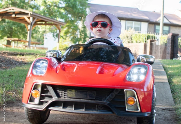 Fototapeta Caucasian baby in a white hat riding in an electric convertible red sports car. child driving a car on a summer day
