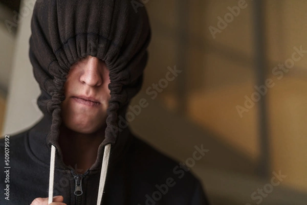 Fototapeta tense teenage guy in a black pulled-down hood covering the upper part of his face. Transitional age and adolescent problems. The teenager wants to hide from everyone.