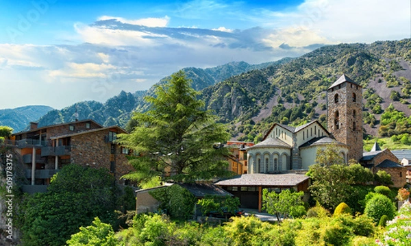 Fototapeta Cityscape of Andorra la Vella city with old houses, ancient Church of Sant Esteve and mountain range against the blue sky on a sunny day. 
Andorra la Vella, the capital of principality of Andorra