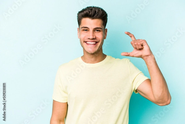 Fototapeta Young caucasian man isolated on blue background holding something little with forefingers, smiling and confident.
