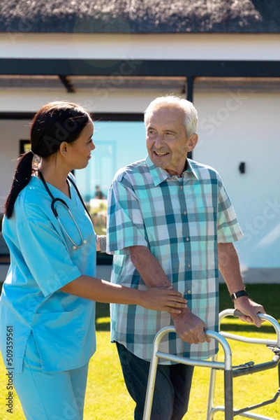 Fototapeta Biracial female doctor helping caucasian senior man in walking with walker in yard on sunny day