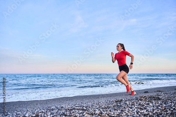 Fototapeta young athletic woman in a red shirt and braid running on the shore of the beach with mountains in the background