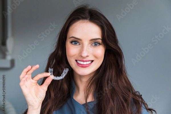 Fototapeta Close-up of orthodontic silicone transparent teeth aligner in female hands. A woman with a perfect charm smile holds a removable night retainer. Bracket for teeth whitening.