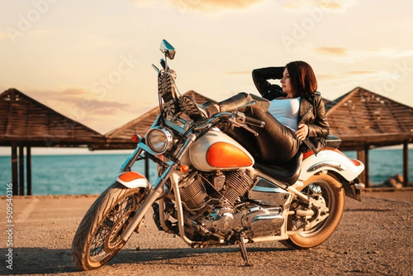 Fototapeta Cool confident adult woman in leather jacket and boots posing lying on motorcycle. Beach umbrellas on the background. The concept of Motorcyclist Day and feminism