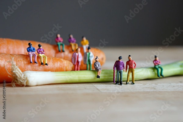Fototapeta Miniature figurine character: people sitting on carrots and spring onions, and some standing on wooden floor, concept of fresh organic vegetables for good health in the market, selected focus