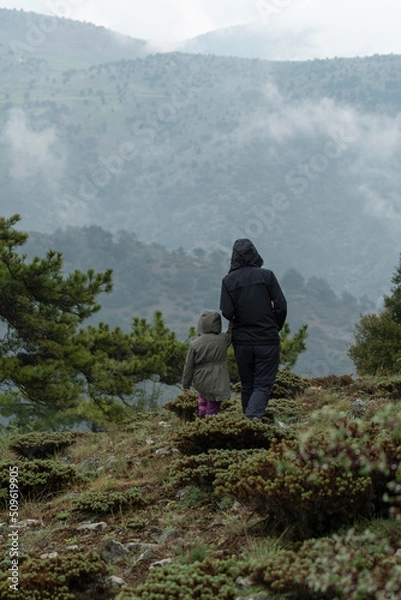 Fototapeta Selective focus shot of mother and daughter standing on mountainside in foggy weather while looking at landscape.