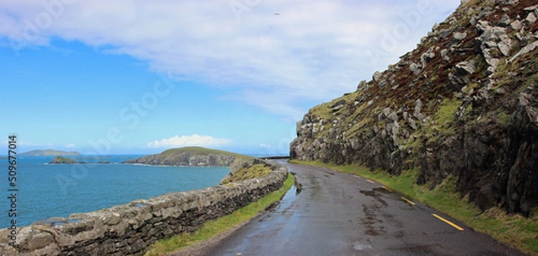 Fototapeta Road between grassy stone slope and sea in western Ireland in blue overcast weather still wet and full of puddles from the rain
