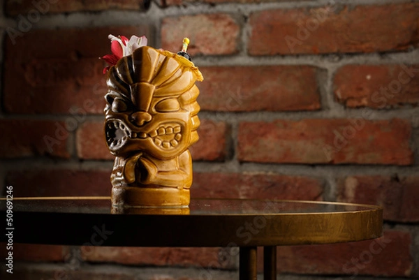 Obraz Tropical cocktail - zombie, served in a tiki style with fruits and flowers. An exotic cocktail in a tiki glass stands on a wooden bar counter in a pub or restaurant, space for text.