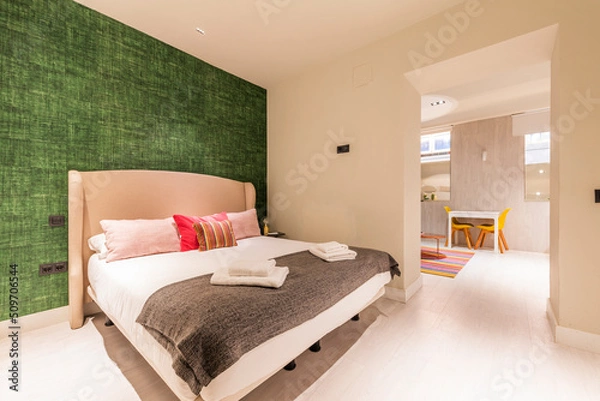 Fototapeta Bedroom with double bed with winged headboard upholstered in fabric, access to living room with white table and light stoneware floor