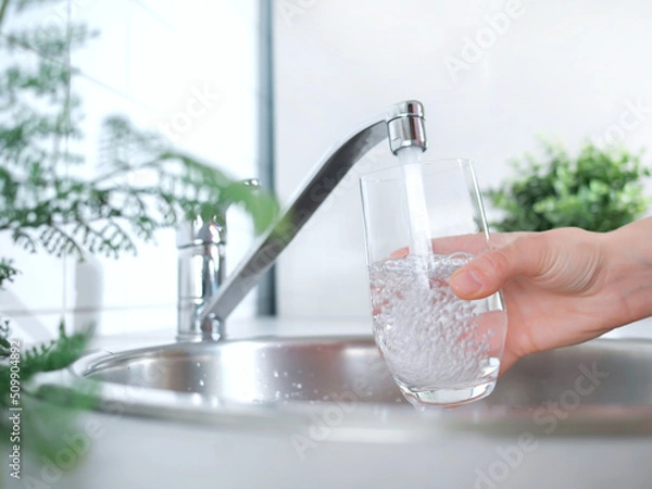 Fototapeta A stream of clean water drink flows into the glass. Woman holding a glass of water under running water from the tap in the kitchen.
