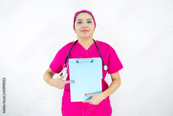 Fototapeta beautiful woman medical assistant, wearing uniform and pink surgical cap, stethoscope, pointing at clipboard with results, on white background