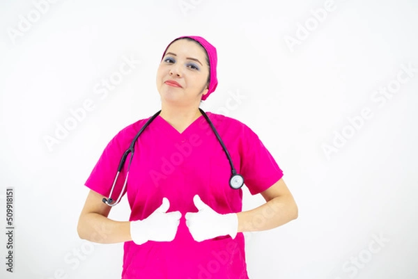 Obraz Beautiful female medical assistant wearing uniform and pink surgical cap, stethoscope, putting latex gloves, on white background