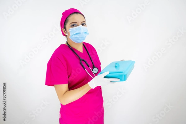 Obraz beautiful female medical assistant wearing face mask, pink uniform and pink surgical cap, stethoscope, preparing instruments case on white background