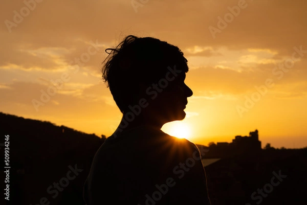 Fototapeta Panoramic silhouette shot of an Indian kid looking at the orange sky during the sunset. Hopeful kid stares at the sun. Kid lost in deep thoughts after looking at the sunset. 