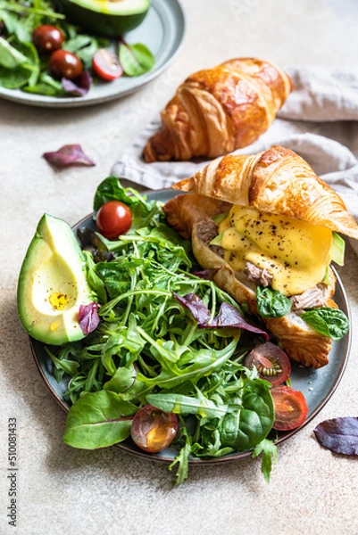 Obraz Croissant with tuna, poached egg, avocado and hollandaise sauce with salad leaves and tomatoes. Croissant sandwich with egg benedict and tuna.