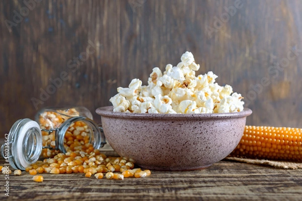 Fototapeta A large bowl of popcorn, corn grains and corncob on a wooden table. Close-up.