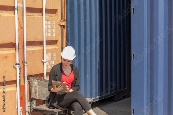 Fototapeta Caucasian female worker uses tablet or smart phone to check stock in a logistic shipyard next to a blue container. Lady in suit with white hard hat uses digital device for her inventory