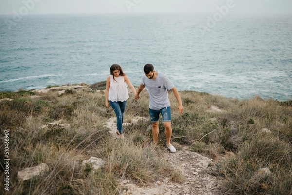 Obraz A Hispanic man and his Latina girlfriend are hiking holding hands in a highland park near the sea in Spain. A couple of tourists are enjoying each other on a date at sunset in Valencia.