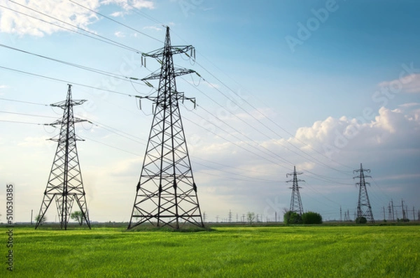 Fototapeta High voltage lines and power pylons in a flat and green agricultural landscape on a sunny day with clouds in the blue sky. Cloudy and rainy. Wheat is growing