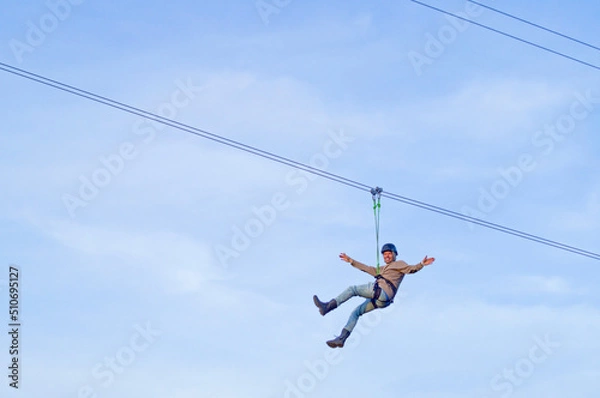 Fototapeta Man practicing zipline under blue sky with clouds