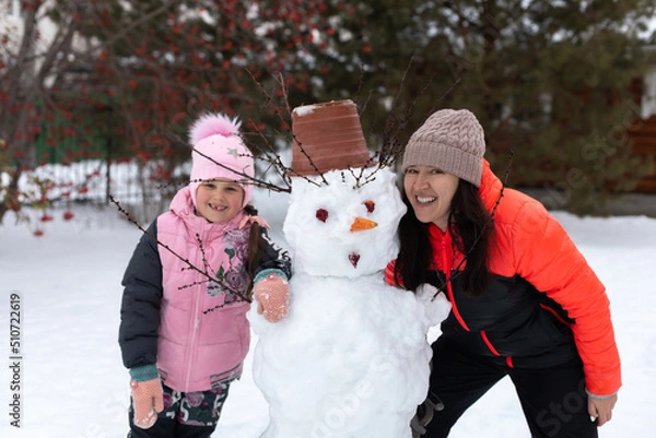 Fototapeta Portrait of little girl and woman with built snowman with snow smiling with teeth in winter while having walk in park with trees in background. Parents spending time with children.