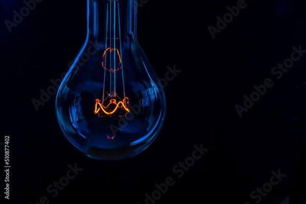 Fototapeta Glowing lamp, tungsten light bulb lit on black background,  close up shot, on wood table,  idea concept,  natural wax candle, Burning candle near a switched off light bulb in complete darkness.