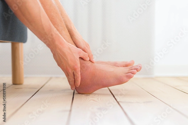 Obraz Ankle sprain. Man hold his sprained ankle sitting on the sofa at home. Sprained and swollen ankle care. Injury.	