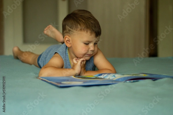 Fototapeta Dressed in casual clothing cheerful boy reads book lying down on a bed in living room.
