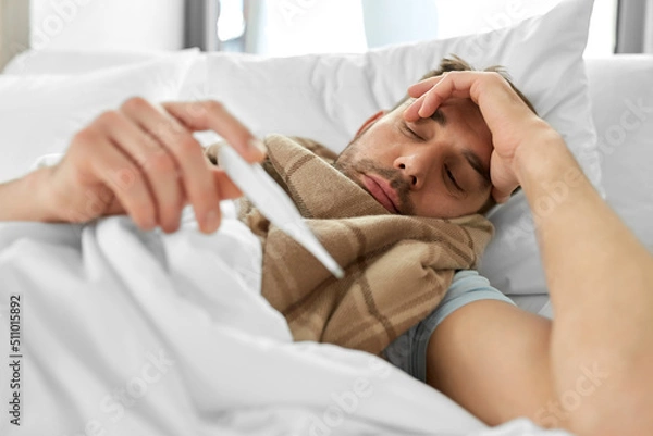 Fototapeta people, health and fever concept - sick man in scarf measuring temperature by thermometer lying in bed at home