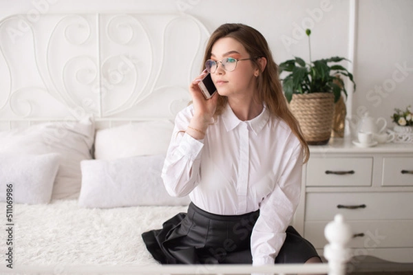 Fototapeta A happy emotional European girl in a white shirt and glasses is talking on the phone. Sitting on a white bed