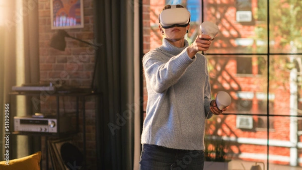 Obraz Young Female Gamer with Short Hair Having Fun and Playing Virtual Reality Game at Home in Her Living Room. She is Wearing a Modern Headset and Using Controllers. Stylish Apartment with Big Window.