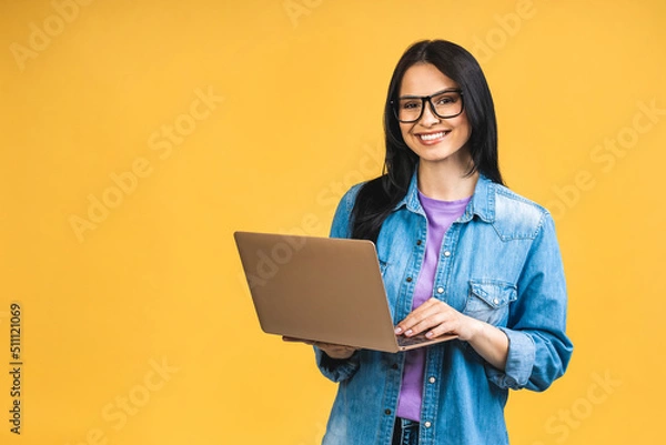 Fototapeta Portrait of happy young beautiful surprised woman with glasses standing with laptop isolated on yellow background. Space for text.